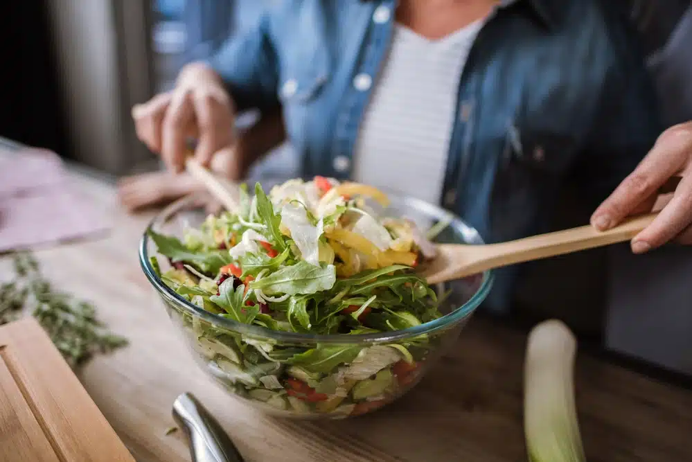 close-up of woman tossing a salad in a glass mixing bowl - nutrition concept
